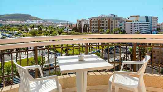 Utsikt fra en balkong med et bord og to stoler, over en by og grøntområder. (AI-generert tekst)