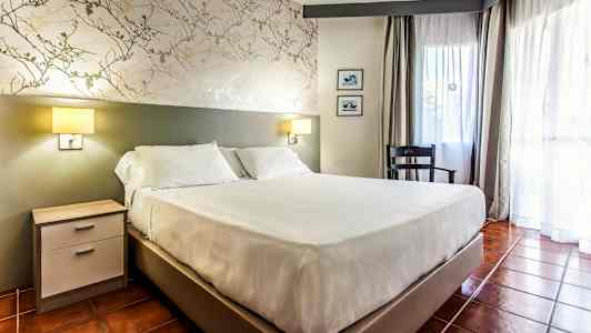 Et rom med en dobbeltseng, nattbord, stol og vindu med gardiner. (AI-generert tekst)