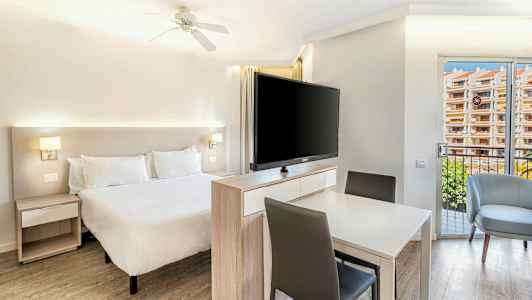 Lyst soverom med en dobbeltseng, et bord med stoler og en stor TV foran sengen. (AI-generert tekst)