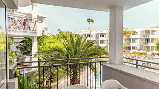 Utsikt fra en balkong mot flere hvitmalte bygninger med frodige palmer og et bassengområde nedenfor. (AI-generert tekst)