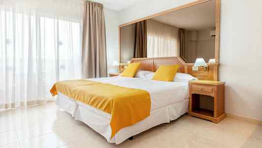 Et hotellrom med en dobbeltseng, hvitt og gult sengetøy. Et stort speil bak sengen og lyse gardiner ved vinduet. (AI-generert tekst)