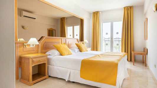 Et hotellrom med en dobbeltseng, hvite laken og gule puter og sengeteppe. Rommet har lyse tremøbler og vinduer med gardiner. (AI-generert tekst)