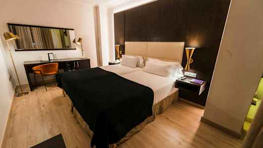 Et hotellrom med en dobbeltseng, mørke og lyse fargetoner på møbler samt en skrivebordskrok med stol og speil. (AI-generert tekst)