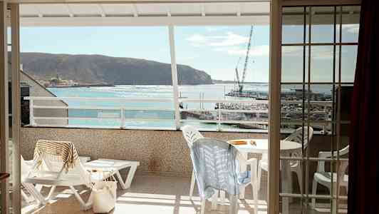 Utsikt fra en balkong mot havet og en liten havn, med utemøbler og solsenger på balkongen. (AI-generert tekst)