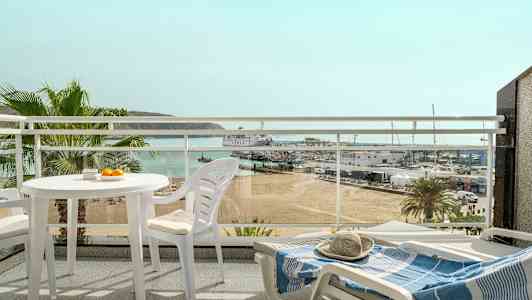 Utsikt fra en balkong med hvite utemøbler, med utsyn over en strand, en småbåthavn og havet i bakgrunnen. (AI-generert tekst)