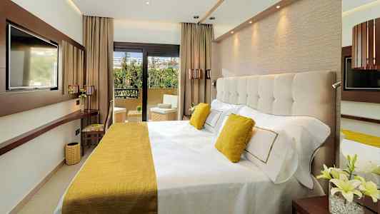 Et moderne hotellrom med en bred seng, innredet med gule detaljer og utsikt mot en balkong. (AI-generert tekst)