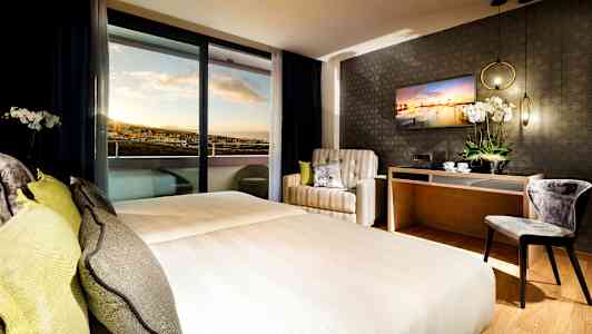 Hotellrom med dobbeltseng, lenestol, skrivebord og utsikt mot solnedgangen. (AI-generert tekst)