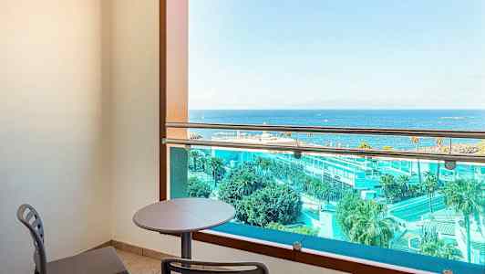 En balkong med bord og stoler, utsikt over havet og hotellområde. (AI-generert tekst)