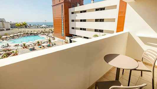 Utsikt fra en balkong mot et bassengområde med solsenger, parasoller og palmer, samt hvite og røde bygninger og havet i bakgrunnen. To stoler og et bord vises på balkongen. (AI-generert tekst)