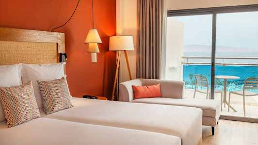 Et hotellrom med dobbeltseng, dekorative puter, en sofa og glassdører som fører til balkongen med utsikt over havet. (AI-generert tekst)