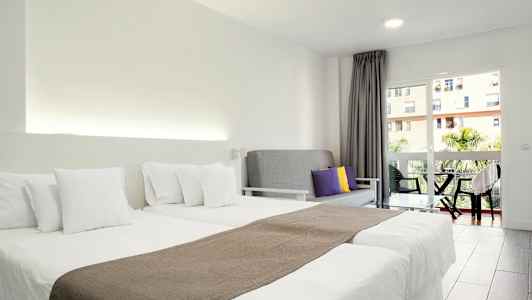 Et hotellrom med to enkeltsenger med hvitt sengetøy, en grå sofa med fargede puter og en balkong med utsikt. (AI-generert tekst)