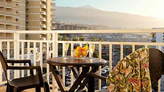 En balkong med et bord, to glass, en skål med frukt og omgitt av en utsikt over omkringliggende bygninger og fjell. (AI-generert tekst)