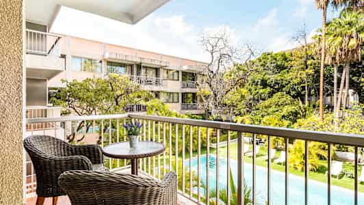 Utsikt fra en balkong med to kurvstoler ved et lite bord over et basseng og grøntområder, samt et boligkompleks i bakgrunnen. (AI-generert tekst)