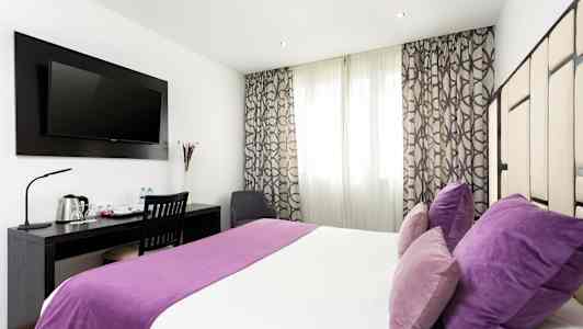 Et hotellrom med dobbeltseng, lilla sengetøy og puter, skrivebord med stol og en veggmontert TV. (AI-generert tekst)