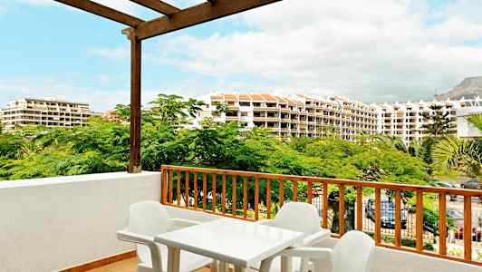 Utsikt fra en balkong med plastbord og stoler, omgitt av grønne trær og bygninger i bakgrunnen. (AI-generert tekst)