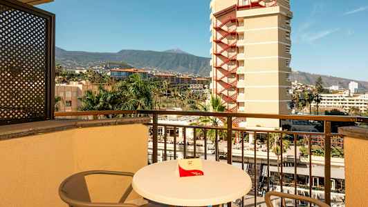 En balkong med bord og stoler, utsikt over omkringliggende bygninger, palmer og fjell i bakgrunnen. (AI-generert tekst)