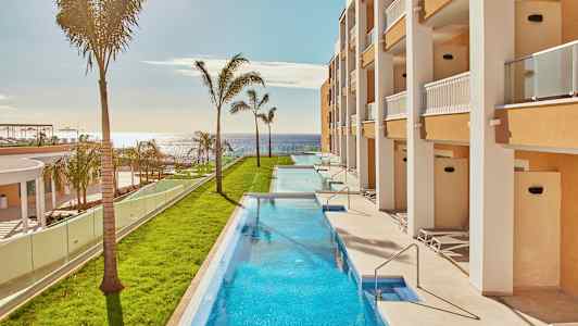 Langsmal basseng omgitt av grøntområder og bygninger med balkonger, med havet i bakgrunnen. (AI-generert tekst)