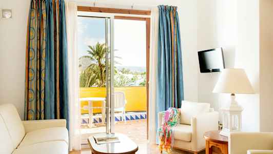 Et lyst oppholdsrom med hvite møbler og åpen balkongdør mot utsikt med palmer. (AI-generert tekst)