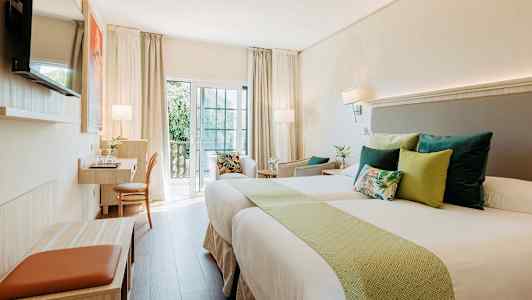 Et lyst hotellrom med to senger, pynteputer og en arbeidsstasjon ved balkongen. (AI-generert tekst)