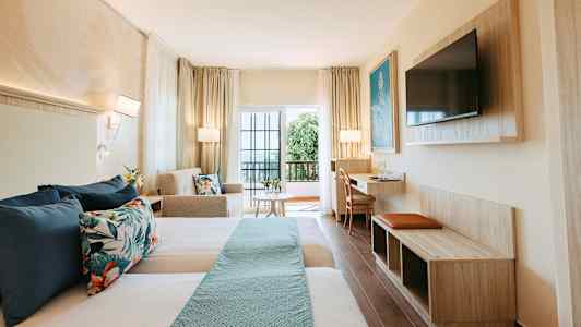 Et hotellrom med to senger, sofa, bord og TV, samt en balkong med glassdører. (AI-generert tekst)