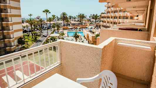 Utsikt fra en balkong mot et basseng og omkringliggende bygninger i et solrikt område. (AI-generert tekst)