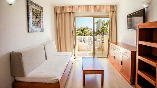 Et møbleret rom med en sofa, et bord, en TV og en hylle. Utgang til en balkong med sittemøbler. (AI-generert tekst)