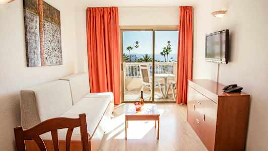 Rom med sofa, bord, TV og balkong med utsikt mot palmer og hav. (AI-generert tekst)