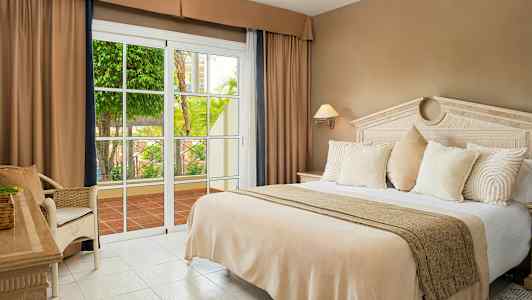 Et hotellrom med en stor seng, beige sengetøy, hvite puter og en glassdør som åpner mot en terrasse med grønne planter. (AI-generert tekst)