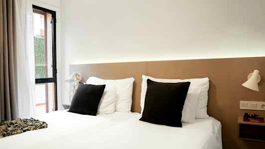 Et hotellrom med to enkeltsenger, hvitt sengetøy og svarte puter, samt lyse nattlamper på veggen. (AI-generert tekst)