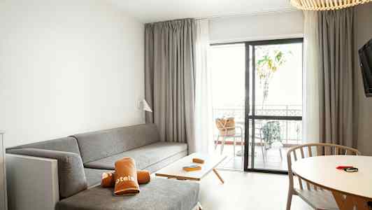 Et hotellrom med en grå sittegruppe, et bord med trestoler og en terrasse gjennom glassdører. (AI-generert tekst)