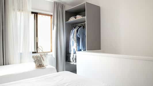 Et rom med to enkeltsenger, gardiner ved vinduet og et åpent skap med klær. (AI-generert tekst)