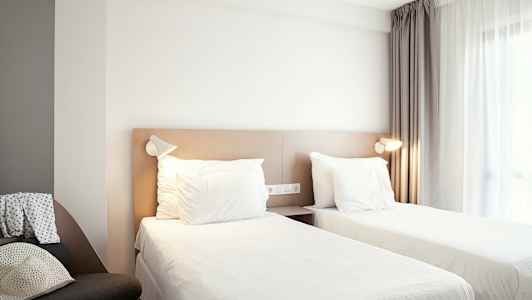 Et hotellrom med to enkeltsenger, et vindu med gardiner og en stol på siden. (AI-generert tekst)