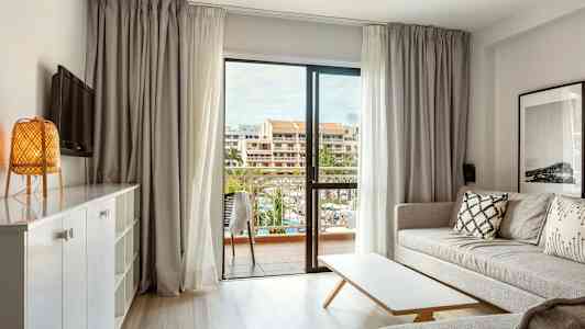 Lyst stue med sofa, bord og utsikt mot en balkong med bygninger i bakgrunnen. (AI-generert tekst)