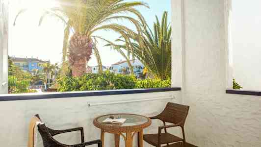 En balkong med to stoler og et bord med en bok. Bak balkongen sees en palme og bygninger. (AI-generert tekst)