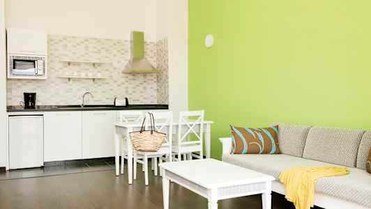 En stue med grønne vegger, en sofa med puter og et bord, samt et tilstøtende kjøkken med hvite skap og apparater. (AI-generert tekst)
