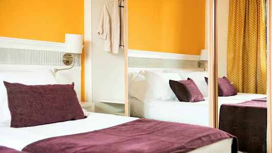 Et hotellrom med en dobbeltseng, lilla puter og oransje vegger. (AI-generert tekst)