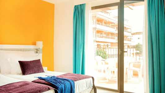 Et hotellrom med en seng, fargerike puter og tekstiler, samt en glassdør som fører til en balkong. (AI-generert tekst)