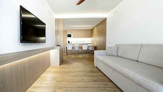 En moderne stue med lys innredning, en grå sofa, en veggmontert TV og et kjøkken i bakgrunnen. (AI-generert tekst)