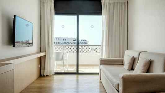 Et lyst og moderne hotellrom med sofa, veggmontert TV og stort vindu med utsikt mot en balkong. (AI-generert tekst)