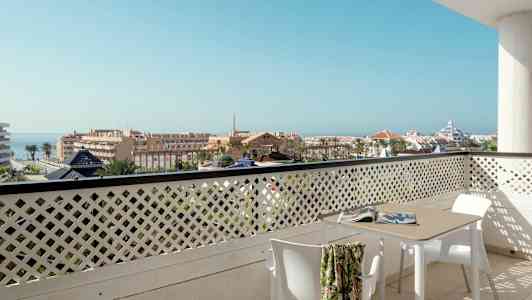 Utsikt fra en balkong med bord og stoler over et bymiljø med havet i bakgrunnen. (AI-generert tekst)