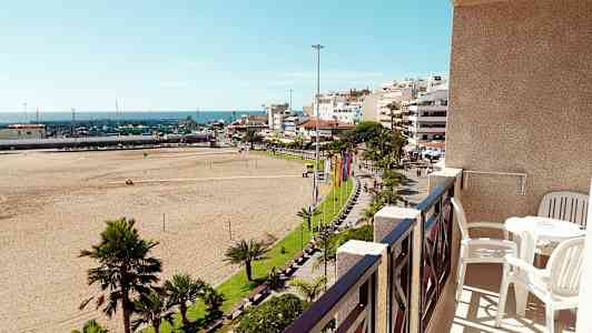Utsikt fra en balkong med bord og stoler mot en sandstrand, frodig park og bymiljø. (AI-generert tekst)