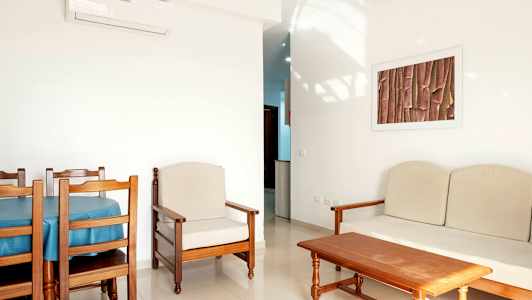 Et rom med lyse vegger og tremøbler, inkludert et spisebord med stoler, en lenestol og en sofa. (AI-generert tekst)