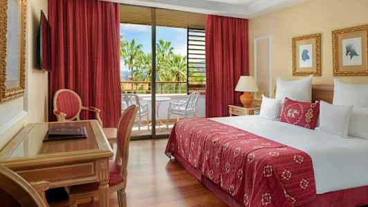 Et hotellrom med en stor seng, sittemøbler og en balkong med utsikt over palmer. (AI-generert tekst)