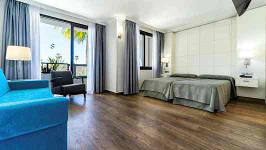 Et hotellrom med to enkeltsenger, en blå sofa, en lenestol og store vinduer med utsikt. (AI-generert tekst)