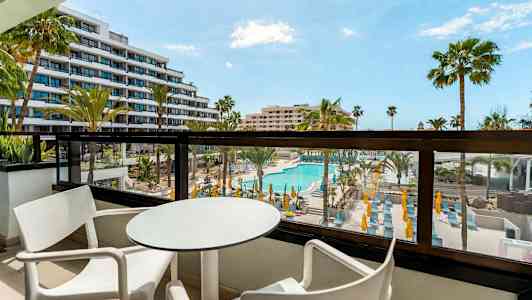 Balkong med bord og stoler, utsikt over bassengområde og omkringliggende bygninger. (AI-generert tekst)