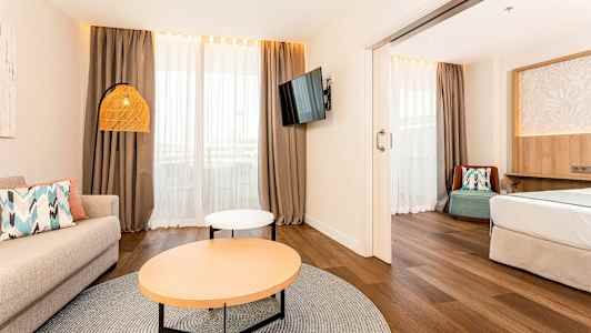 Et lyst hotellrom med en stueområde, sofa, salongbord og en TV på veggen. Sovedelen ligger ved siden av. (AI-generert tekst)