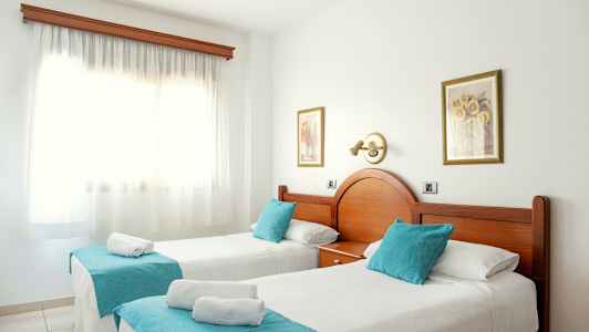 To enkeltsenger med turkise pynteputer og hvitt sengeteppe i et solfylt rom med et stort vindu. (AI-generert tekst)