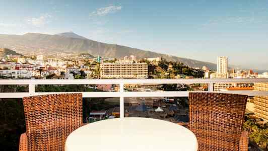 Et bord og to stoler på en balkong med utsikt over en by og et fjell i bakgrunnen. (AI-generert tekst)