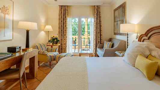 Hotellrom med seng, skrivebord og sittegruppe foran vindu som leder til balkong. (AI-generert tekst)