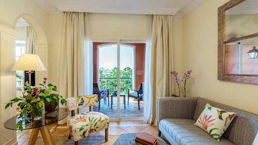 En stue med dekorativ stol, sofa og blomster, med utsikt mot en balkong. (AI-generert tekst)